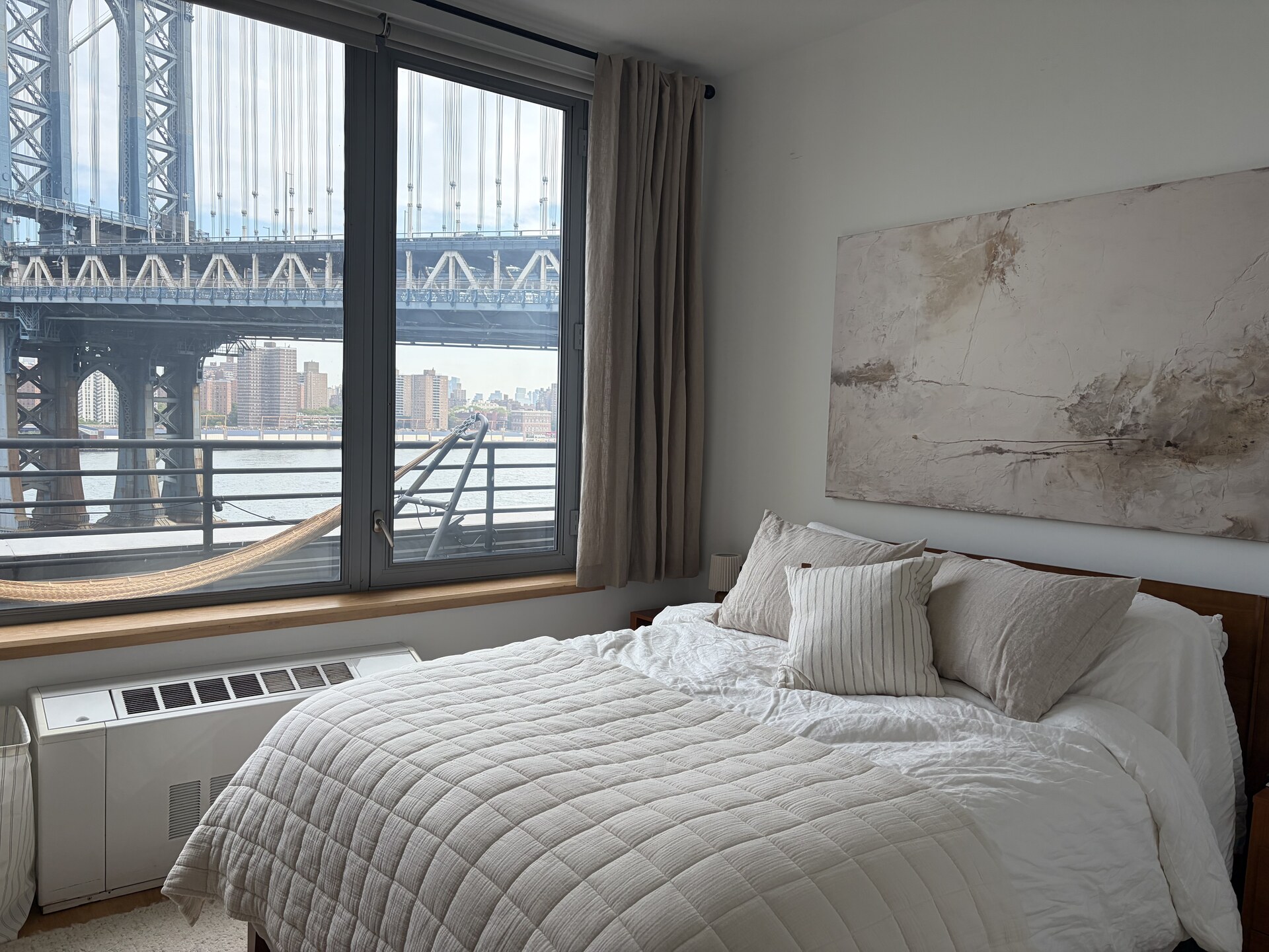A made bed beside a window looking out at the Manhattan Bridge