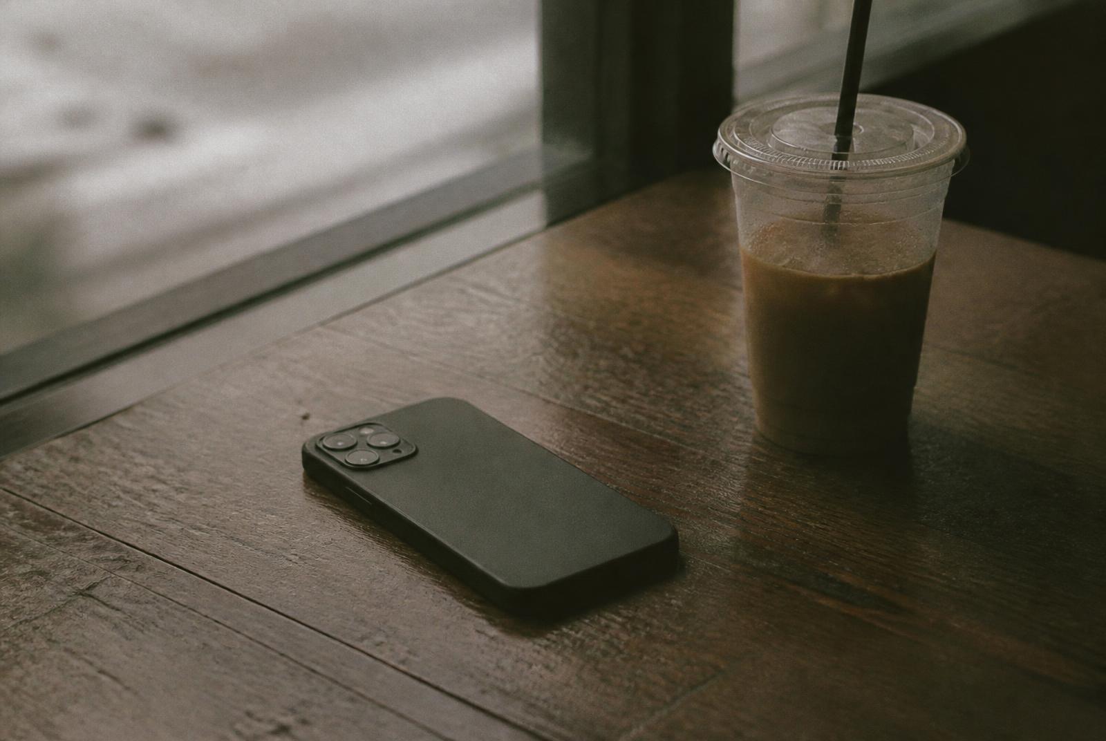 A phone lying face down on a wooden table beside a cold coffee
